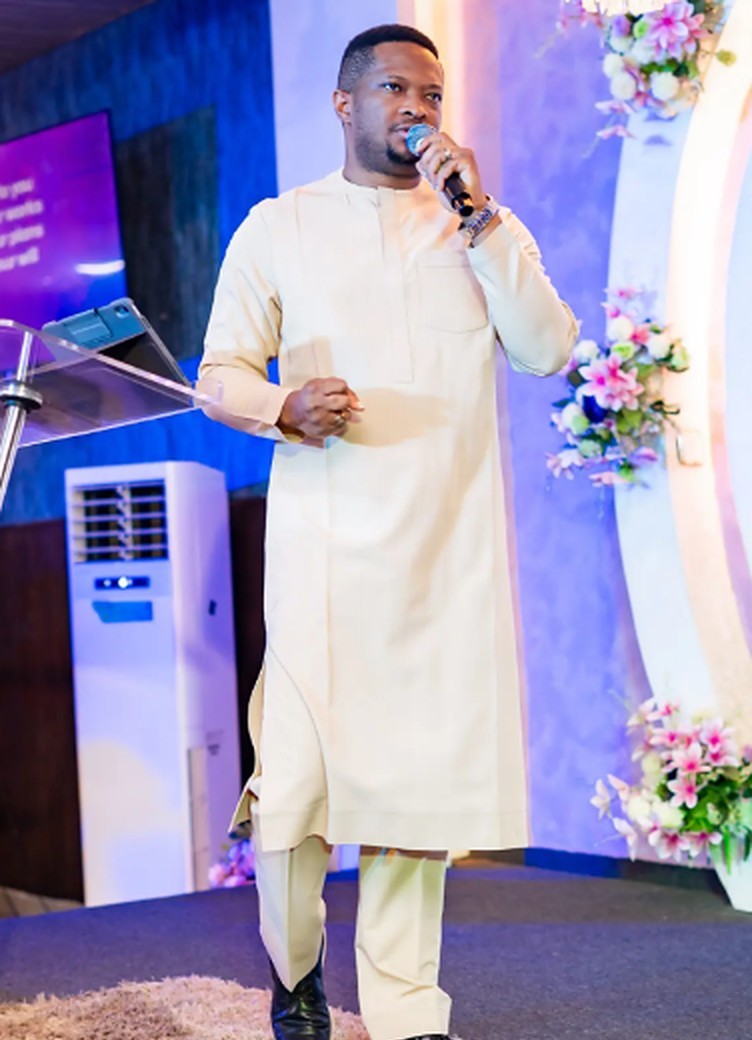 Pastor Godswill Oyor on stage in cream attire, speaking into a microphone